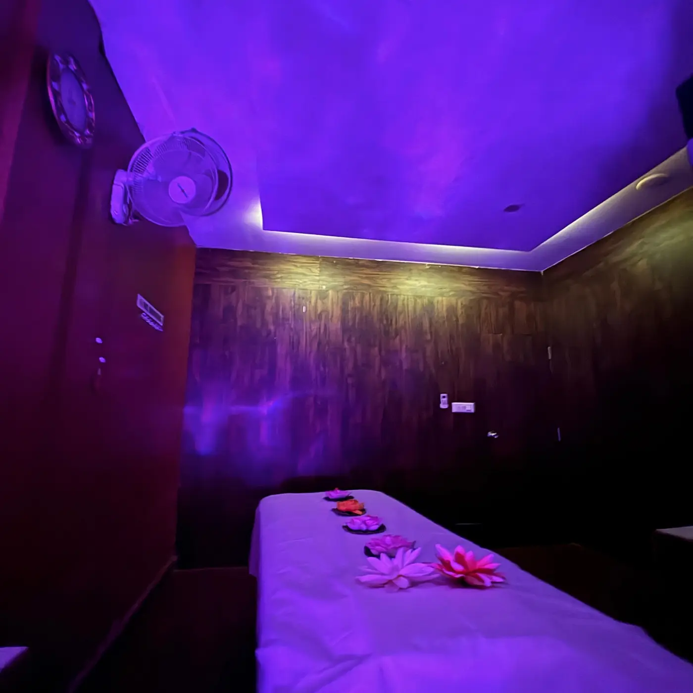 Massage table dressed in fresh white linen with lotus flower accents under soft violet light